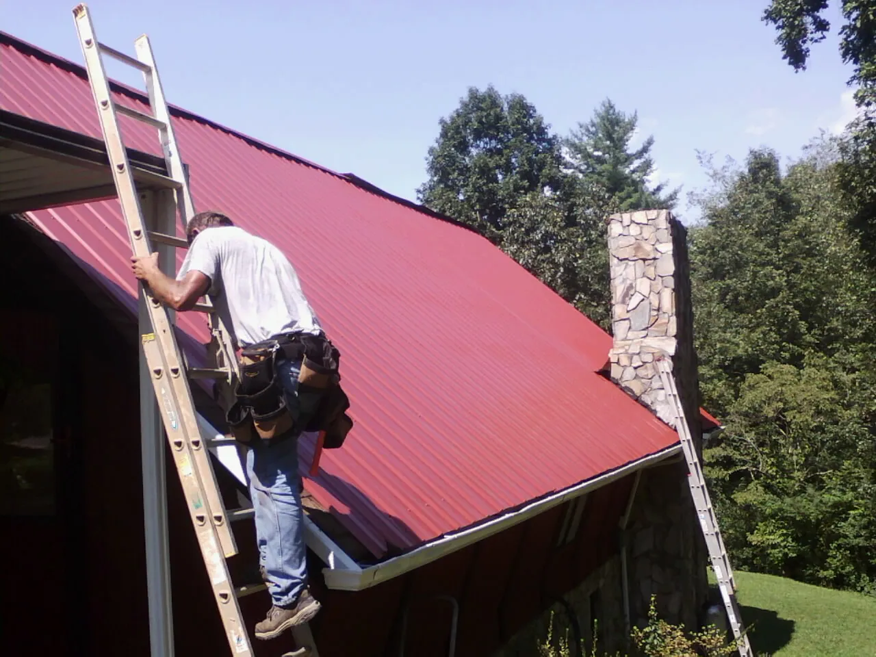Roofer on a ladder beside a red metal roof and stone chimney — Soffit & Fascia Repair by Roofing Brothers in Stewart